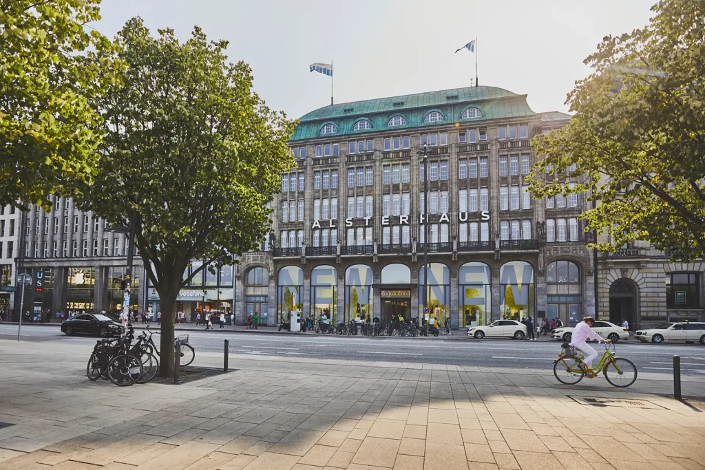 Der Jungfernstieg in Hamburg. Links und rechts sind Bäume zu sehen. Im Hintergrund sieht man das bekannte Alsterhaus.